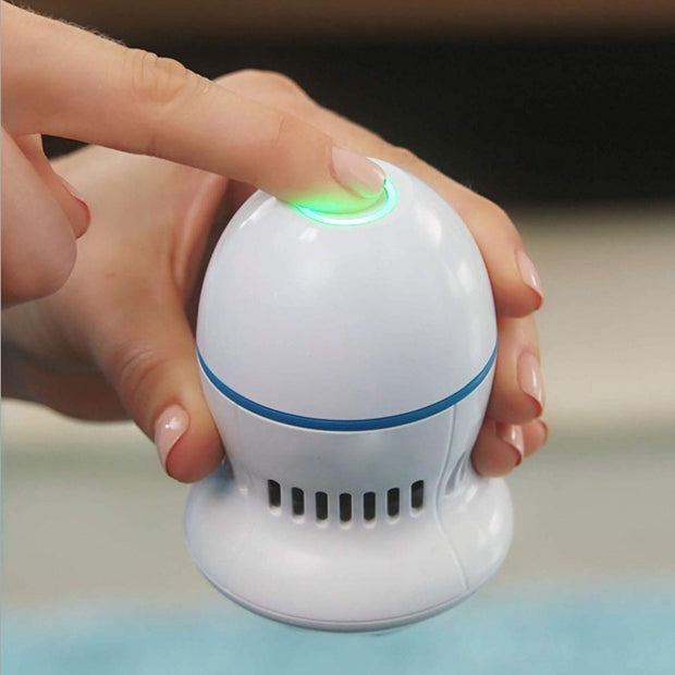 A person presses the glowing green button on top of a white, egg-shaped device designed for callus removal. The device has a vent-like design near its base, and the person's nails are neatly manicured with light pink polish. They are using the ExZachly Perfect Multifunctional Electric Foot File Grinder Machine Dead Skin Callus Remover. The background is blurred.