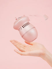 A light pink ExZachly Perfect Multifunctional Electric Foot File Grinder Machine Dead Skin Callus Remover with a cylindrical design, possibly for callus removal or foot massage, hovers above an outstretched hand against a pink background. The device, likely featuring USB charging, has several vents and a button on top, suggesting functionality. Pieces of debris are scattered above the gadget.