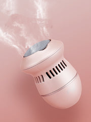 A compact, pink ExZachly Perfect Multifunctional Electric Foot File Grinder Machine Dead Skin Callus Remover emits a misty vapor while charging via USB. The device has a cylindrical body, slotted vents, and a rounded base. It is photographed against a matching pink background, slightly tilted upward as it releases the mist into the air.